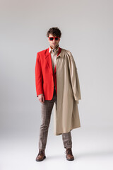 Wall Mural - full length view of fashionable, confident man with trench coat over shoulder looking at camera on grey