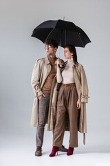 Wall Mural - full length view of stylish couple in autumn clothes standing under umbrella while looking away on grey