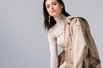 attractive, stylish girl touching collar of trench coat and looking at camera isolated on grey