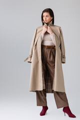 Wall Mural - full length view of fashionable girl touching trench coat and looking away on grey