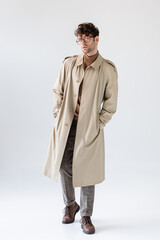 Wall Mural - full length view of fashionable man in trench coat holding hands in pockets while posing on grey
