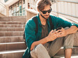 © halayalex - Handsome smiling stylish hipster lambersexual model.Man dressed in white T-shirt.Fashion male sitting on the stairs. Holding, scrolling and texting in his cellphone. Writes sms on smartphone