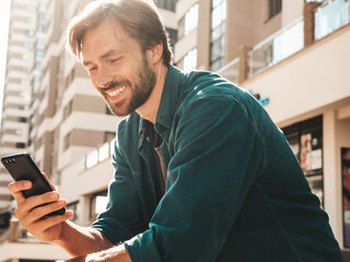 Handsome smiling stylish hipster lambersexual model.Man dressed in white T-shirt.Fashion male posing in the street. Holding, scrolling and texting in his cellphone. Writes sms on smartphone