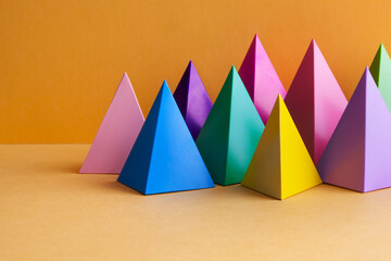 Colorful geometric abstract still life composition. Bright prism pyramid triangle shape figures. Violet yellow blue pink green red color objects, orange background.