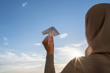 Wall Mural - Silhouette of muslim woman in head scarf launching a paper airplane on blue sunset sky background. Travel concept