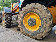 Close-up Of A Large Wheel Of A Yellow Excavator. Dirty Rear Wheel Of An Excavator. Construction Machinery. Detail Of Heavy Tractor Wheel And Tire