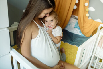 Young pregnant woman sits on a crib in the nursery and hugs her funny curly little daughter 2 years old