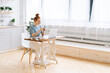 © dikushin - Happy redhead young woman is typing message on cell phone while sitting at the desk in light cozy living room at home office. Girl is looking on display screen of mobile phone. Lady is using cellphone