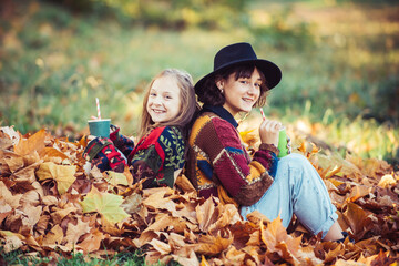 Wall Mural - Happy sisters with autumn yellow leafs. Fallen yellow leaves. Smiling young girl with sister with an orange leaf in hands sitting on autumn foliage in the forest. Sisters enjoying fall nature.