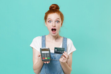 Wall Mural - Amazed young readhead girl in casual denim clothes isolated on blue turquoise background. People lifestyle concept. Hold wireless modern bank payment terminal to process acquire credit card payments.