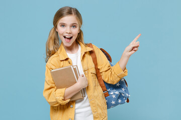 Excited little blonde kid schoolgirl 12-13 years old in yellow jacket backpack isolated on blue background. Childhood lifestyle concept. Education in school. Hold books pointing index finger aside up.