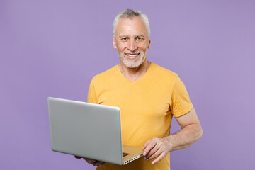 Smiling elderly gray-haired mustache bearded man in casual yellow t-shirt isolated on violet background studio portrait. People lifestyle concept. Mock up copy space. Working on laptop pc computer.