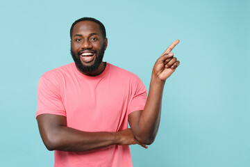 Cheerful young african american man guy in casual pink t-shirt isolated on blue wall background studio portrait. People emotions lifestyle concept. Mock up copy space. Pointing index finger aside up.