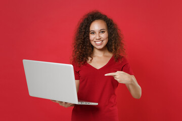 Wall Mural - Smiling young african american woman girl in casual t-shirt posing isolated on red background studio portrait. People lifestyle concept. Mock up copy space. Point index finger on laptop pc computer.