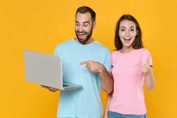 Excited young couple two friends guy girl in blue pink t-shirts isolated on yellow background. People lifestyle concept. Mock up copy space. Pointing index finger on laptop computer, showing thumb up.
