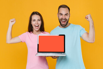 Happy couple friends guy girl in blue pink t-shirts isolated on yellow background. People lifestyle concept. Mock up copy space. Hold laptop pc computer with blank empty screen doing winner gesture.