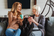© InsideCreativeHouse - Smiling mature mother and adult daughter on sofa in living room talking and drinking tea or coffee