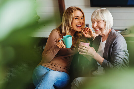 happy senior mother with adult daughter sitting on couch and holding cups with coffee or tea at home