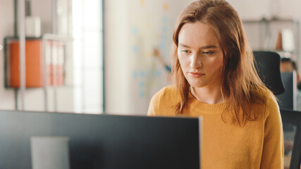Poster - Beautiful and Smart Red Haired Female Specialist Sitting at Her Desk Works on a Desktop Computer. Bright and Modern Open Space Office with Stylish Ergonomic Furniture for Talented Creative People