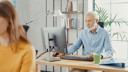 Poster - Handsome Senior Office Employee Sitting at His Desk Works on a Desktop Computer. In the Background Modern Bright Office with Diverse Group of Professionals Working for Growing Startup