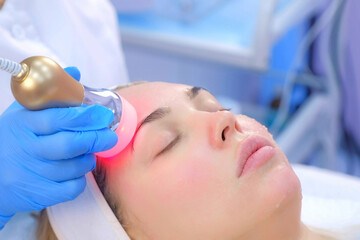 Cosmetologist doctor making phonophoresis procedure on woman face with hyaluronic acid gel, side view. Beautician moving manipula with hot red light on face. Apparatus procedure in cosmetology clinic.