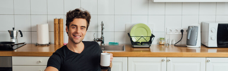 Panoramic shot of handsome man smiling at camera and holding cup of coffee in kitchen