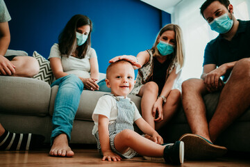 .Young group of friends, accompanied by a small child, having a meeting at home with social distance and surgical masks during the pandemic caused by the covid19