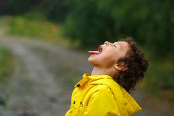 Kid playing in the rain in autumn park. Child jumping in muddy puddle on rainy fall day. Little boy in rain boots and yellow jacket outdoors in heavy shower. Kids waterproof footwear and coat. 