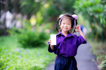 Adorable little asian child girl holding white milk box and drink on green natural background. Kid wearing purple school uniform and sweet smiling in the morning. Children aged 3 - 4 years old.