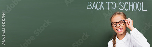 panoramic crop of excited schoolgirl laughing with closed eyes and touching eyeglasses near chalkboard with back to school lettering