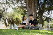© Fernanda - Young Hispanic couple sitting under the tree reading in the park on a sunny day - young people studying with an electronic tablet and a book in the middle of nature - wide shot