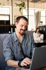 Poster - Smiling handsome young bearded man in denim shirt typing on modern laptop while working in cafe