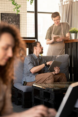 Aufkleber - Young male cafe owner with beard sitting on sofa and pointing at tablet screen while talking to waiter about menu