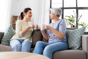 family, generation and people concept - happy smiling senior mother with adult daughter drinking coffee or tea and talking at home