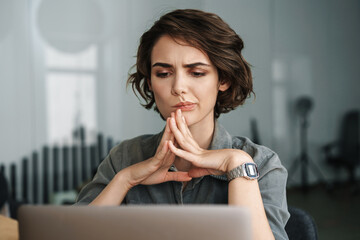 Image of young brooding woman working with laptop while sitting at table