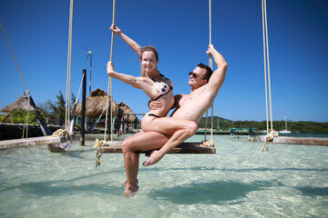 man and woman with beautiful condition body relaxing and posing on swing in clear water in Caribbean sea near resort on the beach