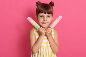 Closeup photo of beautiful little lady two cute knots holding ice creams, licks tongue, wearing white and yellow summer dress isolated pink background,