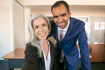 Wall Mural - Happy professional partners posing for photo, smiling and looking at camera. African American successful businessman and Caucasian businesswoman taking selfie. Teamwork and business concept