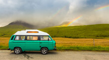 An Old Green Camper Van In The Shadow Of Misty Mountains And A Rainbow