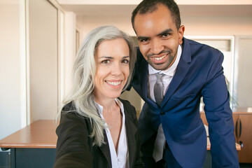 Wall Mural - Cheerful professional partners posing for photo, smiling and looking at camera. African American successful office employer and Caucasian businesswoman taking selfie. Teamwork and business concept