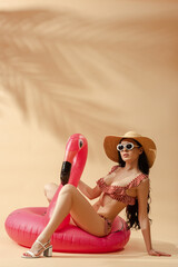 Wall Mural - sexy brunette woman in striped swimsuit, sunglasses and straw hat on inflatable flamingo on beige background