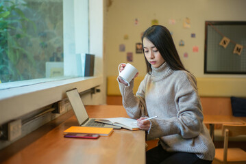 Leinwandbilder - Young business woman while holding a coffee cup and writing on notebook at the cafe.