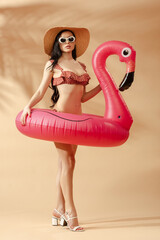 Wall Mural - sexy brunette woman in striped swimsuit, sunglasses and straw hat with inflatable flamingo on beige background