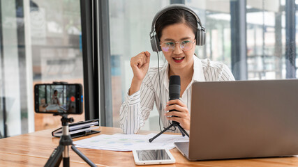 Asian women podcaster podcasting and recording online talk show at studio using headphones, professional microphone and computer laptop on table looking at camera for radio podcast