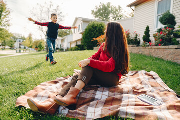 Wall Mural - Elegant woman in a autumn park. Family near house. Mother sitting on a plaid with her little son.