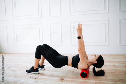 Asian female sportswoman does an exercise to strengthen the muscles of the back and hips with the help of the massage roller for the fascia.