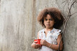 © LIGHTFIELD STUDIOS - Poor african american child looking at camera while holding metal cup near concrete wall in slum