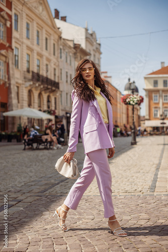 Outdoor full-length fashion portrait of elegant woman wearing lilac suit: blazer, trousers, strap sandals, holding trendy big white leather pouch handbag, walking in street of European city
