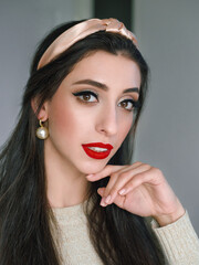 Wall Mural - Close up headshot portrait of young natural looking casual beautiful brunette woman with trendy makeup and vibrant red lips in hair hoop and pearl earrings domestic room background