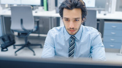 Wall Mural - Handsome Motivated Office Worker Uses Desktop Computer, Typing on a Keyboard. Confidentand Young Businessman Works in the Big Bright Office with Team of Professionals